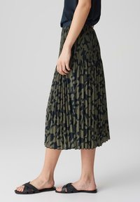 Gonna midi plissettata di un verde scuro con un motivo astratto nero. Indossata con sandali neri con cinturini, che mostrano un design fluido e una trama del tessuto.