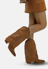 Botas de cowboy de ante marrón con puntas afiladas y costuras decorativas. Altura hasta la mitad de la pantorrilla con tejido texturizado y un tacón cuadrado.