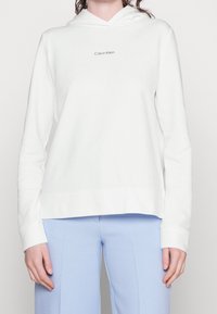 Sudadera blanca con capucha, de corte holgado, mangas largas y un pequeño logo "Calvin Klein" en el pecho. Textura de tela suave.