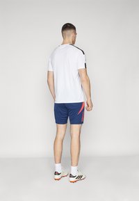 Biela košeľa s krátkym rukávom a čiernymi akcentmi, v kombinácii s námorníckymi šortkami s červeným diagonálnym pruhom. Nosí sa s bielymi a šedými futbalovými kopačkami.