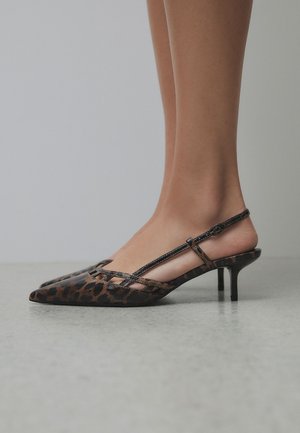 Stopy w brązowych szpilkach typu slingback z panterkowym wzorem, w szpiczastym noskiem i niskim obcasem typu szpilka, na jasnoszarej podłodze.