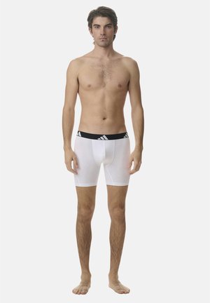 Mann steht barfuß mit Blick nach vorne, trägt weiße Boxershorts mit einem schwarzen Bund, auf dem ein weißes Adidas-Logo zu sehen ist.