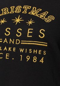 Tissu noir avec texte brodé en or : "NOËL," étoiles, "BISOUS ET SOUHAITS DEPUIS 1984."