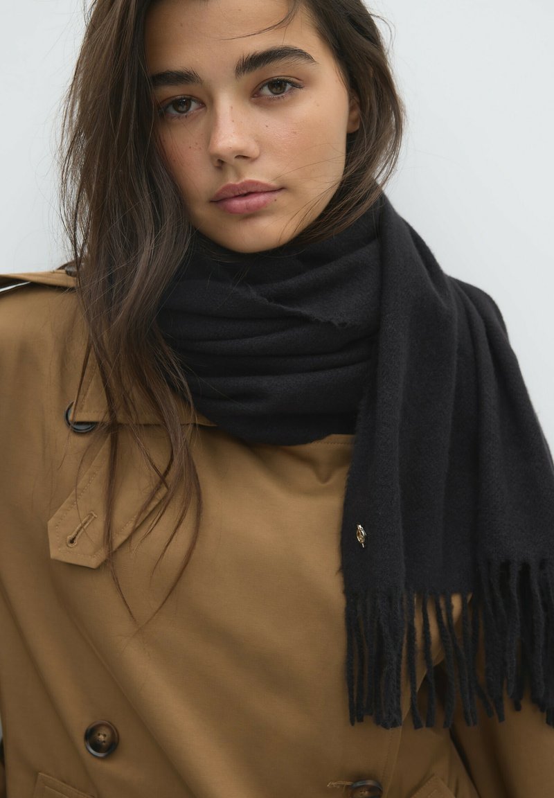 Jeune femme aux longs cheveux bruns portant un manteau beige et une écharpe noire, regardant directement avec une expression neutre sur un fond uni.