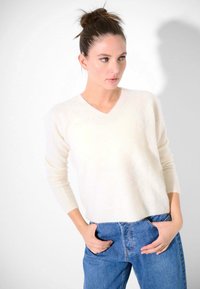 Maglione beige a V, realizzato in un morbido tessuto testurizzato, con maniche lunghe, abbinato a jeans blu a vita alta. Design semplice e classico.