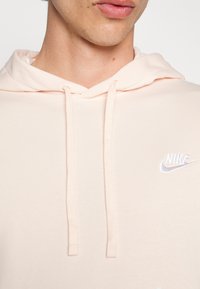 Ljusrosa bomullshoodie med dragsko, har en liten vit Nike-logotyp på bröstet, och visar en mjuk, slät textur.