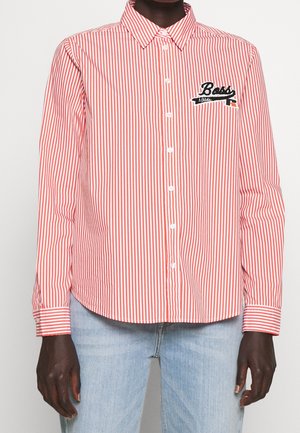 Person iført en rød og hvid skjorte med lodrette striber og knapper, med "Boss Athletic" broderet på venstre bryst, kombineret med lyseblå jeans.