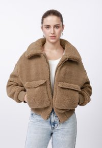 Fiatal nő túlméretezett barna polárdzsekit visel nagy zsebekkel, fehér felsővel és világoskék farmerrel, egyszínű fehér háttér előtt.