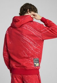 Hoodie rouge avec motif éclaboussé blanc, capuche réglable avec cordon, poignets côtelés et un patch étiquette à l'arrière près de l'ourlet.
