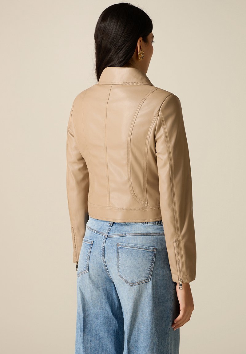Vrouw die een beige, nauwsluitend leren jack draagt met ritssluiting bij de manchetten en lichtblauwe, hoog taille jeans, gezien van achteren tegen een effen achtergrond.