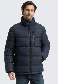 Jūras zila pūkā fūza jaka ar horizontālu quiltēšanu, augstu apkakli, priekšējo rāvējslēdzēju un sānu kabatām. Izgatavota no gludas, izolējošas vielas.