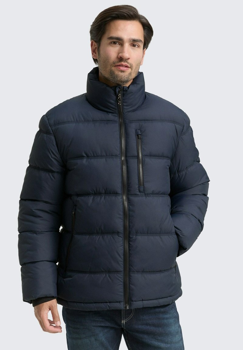 Jūras zila pūkā fūza jaka ar horizontālu quiltēšanu, augstu apkakli, priekšējo rāvējslēdzēju un sānu kabatām. Izgatavota no gludas, izolējošas vielas.