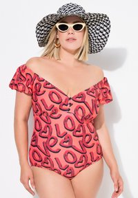 Rosa baddräkt i ett stycke med volangärmar utan axlar, med ett svart hjärttryck. Tillbehör inkluderar en svart och vit mönstrad bredbrättad hatt och solglasögon.
