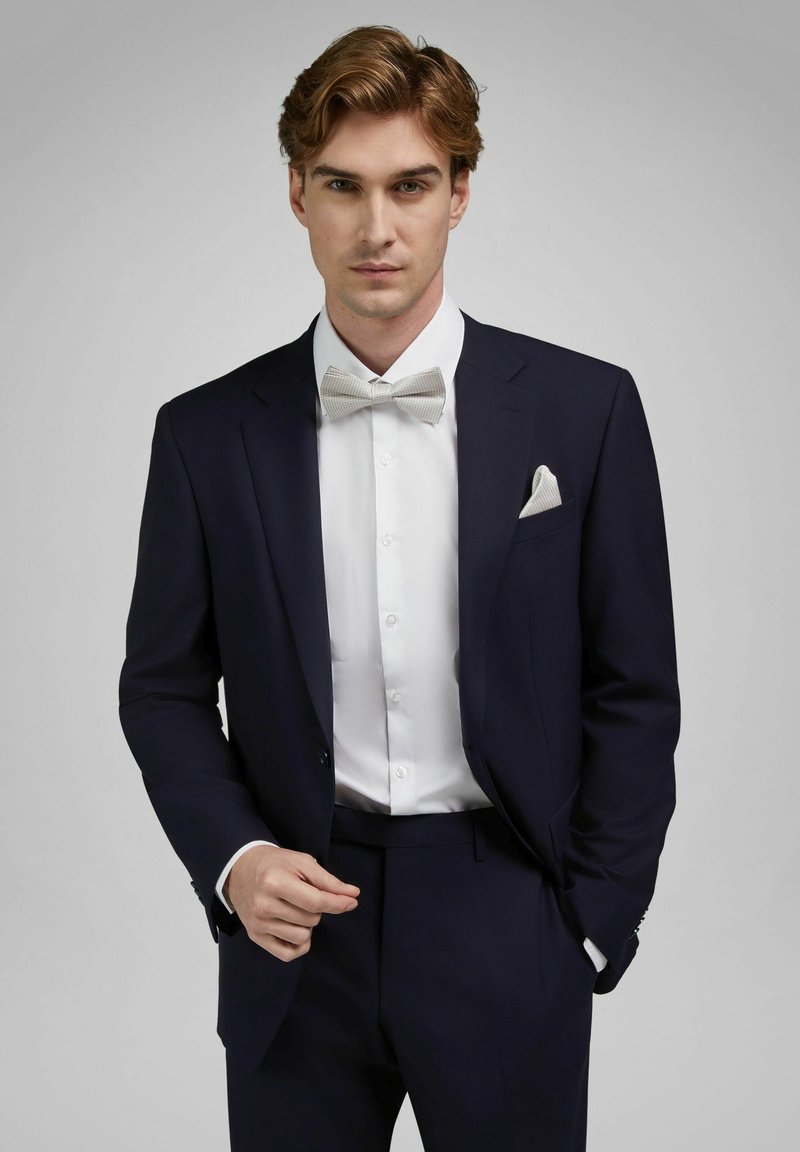 Young man in dark navy suit with white dress shirt, light gray bow tie, and matching pocket square, posing against light gray background.