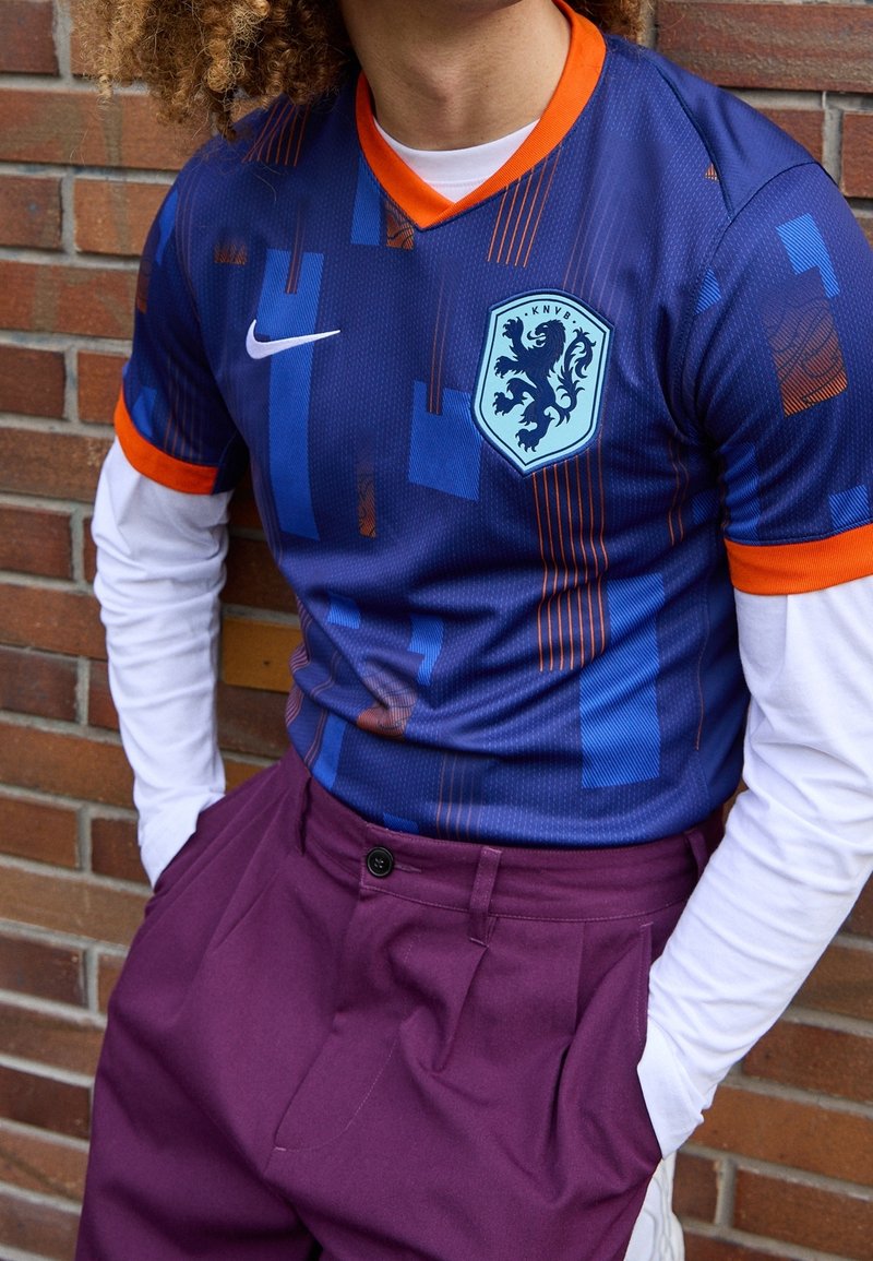 Maillot de football bleu avec des motifs géométriques, des accents orange et un emblème de lion. Chemise blanche à manches longues en dessous, associée à un pantalon bordeaux.