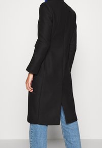 Abrigo negro ajustado con una silueta larga, textura suave, abertura lateral y mangas tres cuartos, combinado con jeans de mezclilla azul claro.