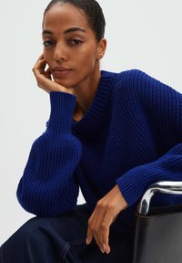 Pull bleu tricoté avec une large texture côtelée, doté d'un col rond et de manches volumineuses. Le modèle repose son menton sur sa main. Jean foncé visible.