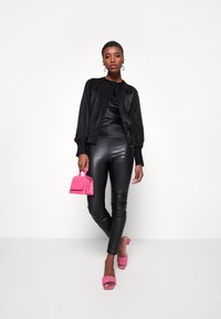 Veste bomber en satin noir avec un col froncé, accompagnée de leggings noirs brillants. Accentuée par un petit sac à main en cuir crocodile rose.