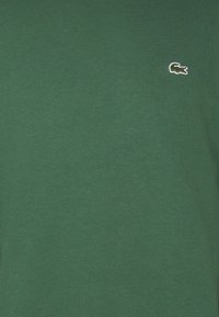 Zelené bavlnen�é tričko s okrúhlym výstrihom a krátkymi rukávmi. Na ľavej strane hrude má malé biele logo Lacoste vyšívané.