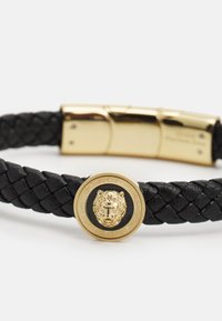 Black braided leather bracelet featuring a gold-tone metal clasp and a circular charm with a lion head emblem in the center.