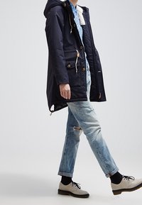 Marinblå parkas med huva, knappar framtill och två sidofickor, kombinerad med ljusblå slitna jeans och grå snörskor.