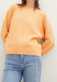 Pomarańczowy prążkowany sweter z okrągłym dekoltem i opuszczonymi ramionami, zestawiony z jasnoniebieskimi dżinsami. Teksturowana tkanina i luźny fason.