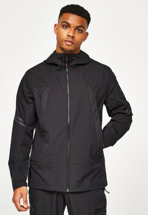 Ung mand iført en sort hættejakke med lynlås og sorte cargobukser, stående foran en ensartet lys baggrund.