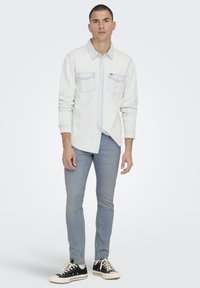 Camisa de mezclilla azul claro con dos bolsillos en el pecho, combinada con jeans de mezclilla claros y zapatillas negras. Textura suave, diseño de botones.