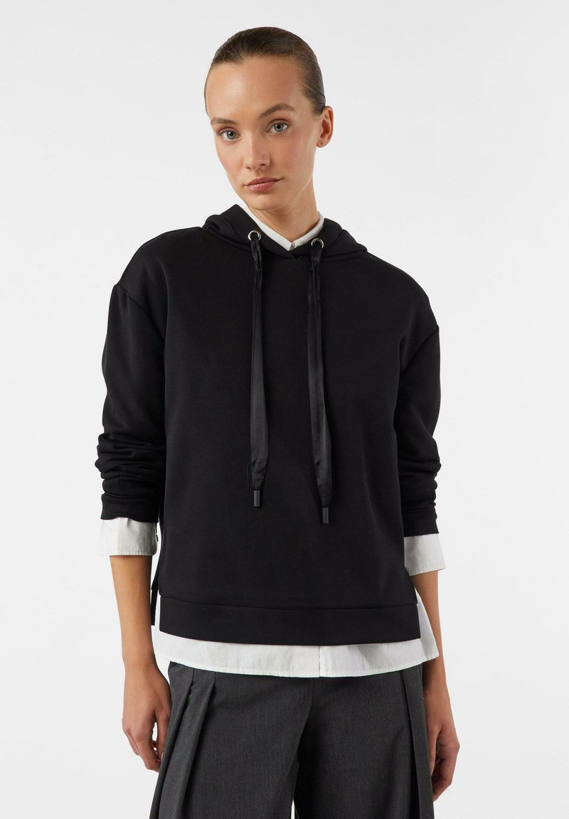 Czarna bluza z kapturem z satynowymi sznurkami, ściągaczami na rękawach i luźnym krojem, noszona na białej koszuli. Miękka faktura tkaniny.