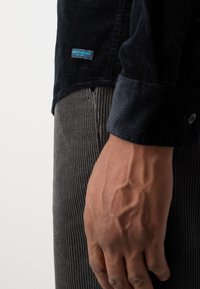 Camicia in velluto a coste scuro con orlo curvo, con un'etichetta blu. Abbinata a pantaloni in velluto a coste grigio testurizzato. Mano visibile che afferra il tessuto.