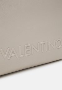 Ljus beige strukturerad läderhandväska med präglad "VALENTINO"-logotyp. Slät yta med minimalistisk design och synliga sömnadsdetaljer.