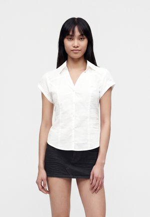Femme aux longs cheveux noirs portant une chemise blanche à manches courtes boutonnée et une mini-jupe en denim noire, debout devant un fond uni.