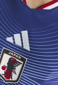 Blå tröja med texturerad tyg, med vita ränder, en Adidas-logga och en Japan Football Association-logotyp längst ner.