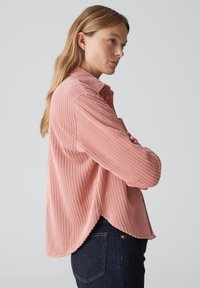 Pink ribbed long-sleeve shirt with a collared neckline and button accents. Features a relaxed fit and rounded hem, paired with dark jeans.