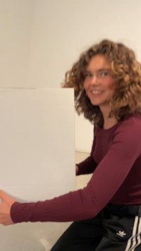 Caja cuadrada blanca sostenida por una persona que viste una camisa de manga larga burdeos y pantalones negros con rayas blancas, en un interior con paredes de colores neutros.