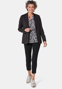 Zwarte blazer over een zwart-witte blouse met patroon, gecombineerd met cropped zwarte broek en grijze sneakers met witte accenten.
