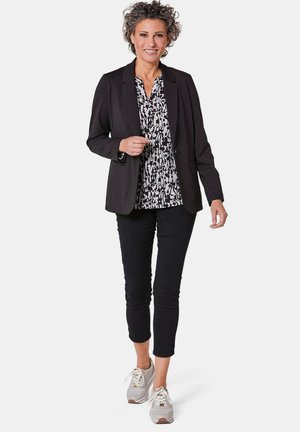 Zwarte blazer over een zwart-witte blouse met patroon, gecombineerd met cropped zwarte broek en grijze sneakers met witte accenten.