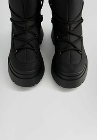 Svarta vattentäta stövlar med vadderad överdel, tjock gummisula och korsade snördesign för en säker passform. Texturerad yta.