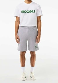 T-shirt blanche avec le texte "CROCODILE" en vert, assortie à un short en molleton gris avec le texte "GAME. SET. MATCH!" en vert, le tout complété par des chaussettes et des chaussures blanches.