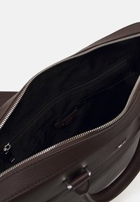 Tummanruskea nahkabag, jonka ylävetoketju on auki, paljastaen mustan kankaan sisäosan, jossa on sisäinen vetoketjullinen tasku ja kohokuvioitu brändilappu.