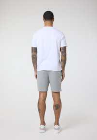 Baltas medvilninis marškinėliai su trumpomis rankovėmis, derinami su pilkais medvilniniais šortais. Asmuo vilki baltus sportinius batelius, ant kojų matomi tatuiruotės.