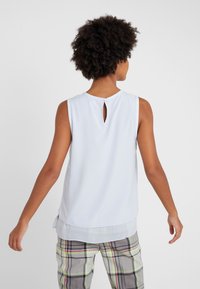 Kvinna med lockigt hår som bär en ärmlös vit blus med nyckelhålsdetalj på ryggen och rutiga byxor med neoninslag, stående mot en enhetlig bakgrund.