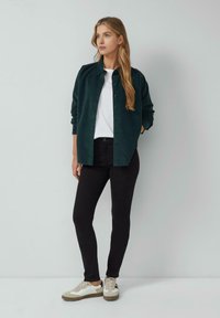 Donkergroen corduroy overhemd over een wit T-shirt, gecombineerd met zwarte jeans en beige sneakers, tegen een neutrale achtergrond.