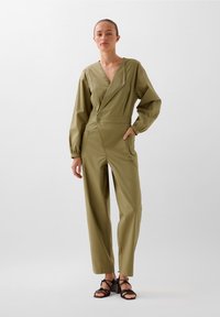 Oliven grøn jumpsuit med en dyb V-hals, lange ærmer, indsnævret talje og brede ben. Knapdetalje ved taljen; båret med remsandaler.