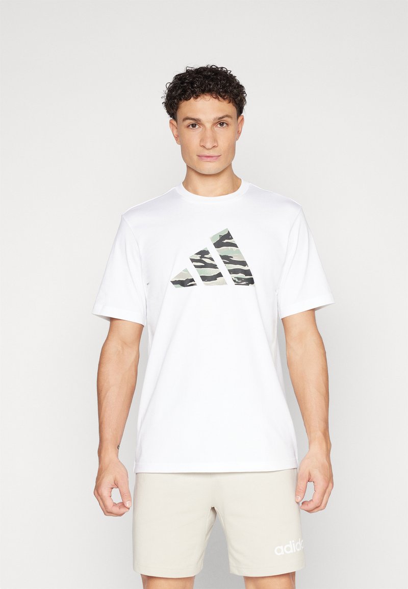 Bijela kratka majica s kratkim rukavima s zelenim kamuflažnim Adidas logom na prsima, napravljena od mekog materijala. U kombinaciji s bež šortsevima.