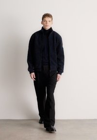 Tumesinine tekstuuriga tõmblukuga bomberjakk musta kaelusega kukla all oleva pluusi peal, kombineeritud mustade pükste ja mustade kingadega, modell kõnnib neutraalsel taustal.