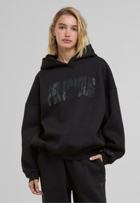 Schwarzer, übergroßer Hoodie aus weichem Stoff, mit einem gewölbten "DELICIOUS" Grafik in glänzenden Akzenten auf der Vorderseite. Doppellagige Kapuze und Kängurutasche.