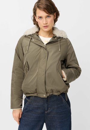 Young person wearing a short olive green jacket with a white faux fur collar and dark blue jeans, standing with one hand in jacket pocket.