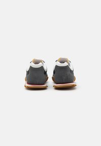 Vista trasera de zapatillas grises y blancas con una parte superior de ante, suela de goma y acentos amarillos en la lengüeta del talón.