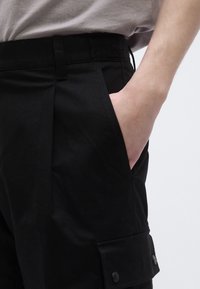 Mustad kaubapüksid, millel on lame esiosa, küljetasku ja nupuga klapiga detail. Valmistatud siledast kangast minimalistliku disainiga.